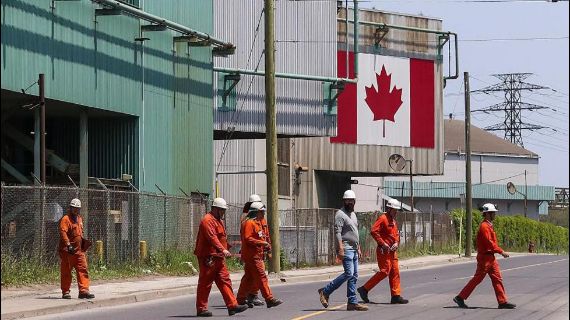 Workers in orange jumpsuits and white hard hats walk in front of a building with the Canadian flag on it